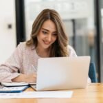 Une Femme Est Assise à Un Bureau, Souriant Tout En Travaillant Sur Un Ordinateur Portable. Des Papiers, Un Presse Papiers Et Une Plante En Pot Sont Posés Sur La Table.