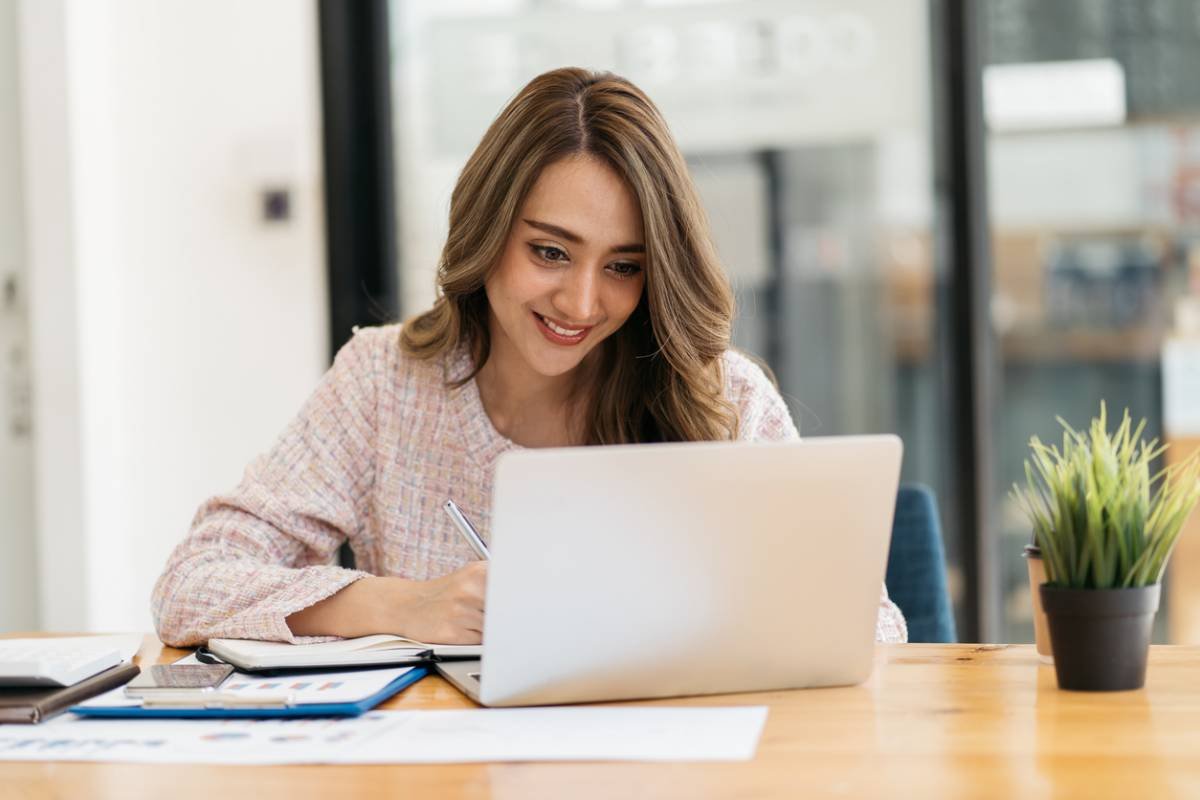 Une Femme Est Assise à Un Bureau, Souriant Tout En Travaillant Sur Un Ordinateur Portable. Des Papiers, Un Presse Papiers Et Une Plante En Pot Sont Posés Sur La Table.