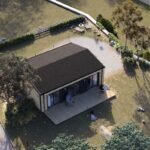 Une Petite Maison Moderne Avec Un Toit Sombre, Une Terrasse En Bois Et Des Sièges Extérieurs, Entourée D'arbres, De Buissons Et D'un Chemin De Gravier à L'intérieur D'une Cour Herbeuse Clôturée.