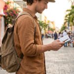 Un homme avec sac à dos et chapeau lit une carte dans une rue pavée bordée de maisons colorées et de passants.