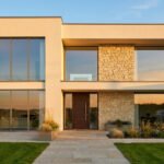 Façade d'une maison moderne beige et pierre, grandes baies vitrées, porte en bois foncé. Chemin pavé et jardin soigné au soleil couchant.