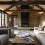 Salon de grange rénovée avec murs en pierre, poutres apparentes, cheminée moderne et grandes baies vitrées. Plans d'architecte sur la table.