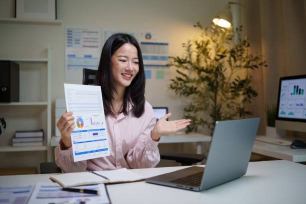 Une Femme Est Assise à Un Bureau Et Tient Un Document Contenant Des Tableaux Et Des Graphiques, Devant Un Ordinateur Portable Dans Un Environnement De Bureau.