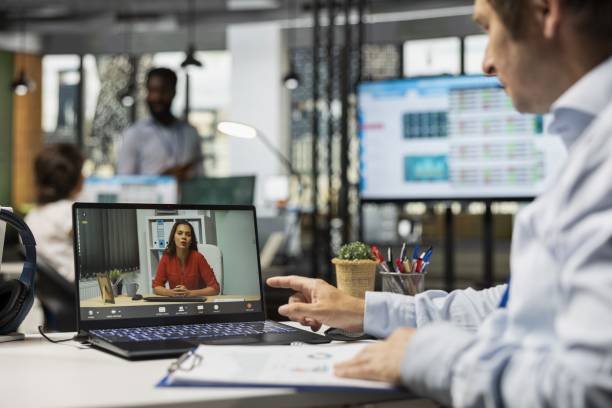 Un Homme Est Assis à Un Bureau Et Participe à Un Appel Vidéo Sur Son Ordinateur Portable Avec Une Femme, Tandis Que D'autres Personnes Travaillent En Arrière Plan.