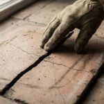 Une main gantée inspecte une profonde fissure traversant un carreau de carrelage ancien au sol, près d'une fenêtre.