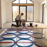Un artisan installe un chauffage au sol dans une pièce en rénovation, avec des tuyaux bleus et rouges raccordés à un collecteur.