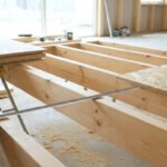 Deux ouvriers mesurent l'entraxe des solives en bois avant la pose de panneaux OSB pour un plancher. Gants bleus visibles.