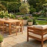 Mobilier de jardin en bois clair (table, chaises, canapé) sur un patio en pierre, au milieu d'un jardin luxuriant avec oliviers et fleurs.
