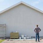Un homme observe le grand pignon gris d'une maison, avec échafaudage et pots de peinture au sol sous un ciel bleu.