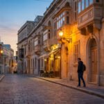 Vue d'une ruelle pavée maltaise au crépuscule, bâtiments en pierre avec balcons éclairés, un homme marche.